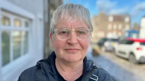 Margaret Rae has short grey hair and is wearing glasses and a dark jacket. She is standing in a street.