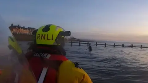 The back of an RNLI crew member in a bright yellow helmet looking out to shallow sea water where a boy is waist deep in water. You can see a town promenade in the background and low evening light.
