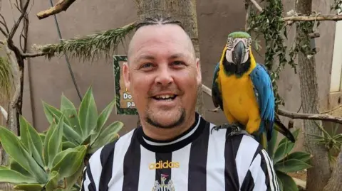 Anthony Robson is wearing a black and white Newcastle top with a parrot on his shoulder. He is smiling to the camera and has a chin beard. 