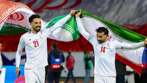 Iran's players Omid Norafkan and Saman Ghoddos celebrate after a qualifying match. They are smiling and dressed in white football kits. One of them is waving an Iranian flag above his head.