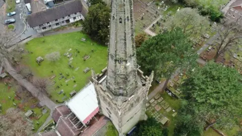 A church tower viewed from above with a large steeple on top. Below is the green graveyard and a black and white timbered building