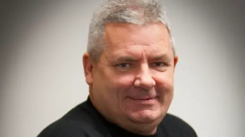 Ollie Green has grey hair, is smiling at the camera  and is wearing a black t-shirt.
