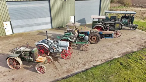 Cheffins An aerial shot of seven vintage tractors, all lined up in front of a warehouse-type building