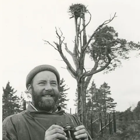 RSPB Scotland In a black and white photograph is a conservationist, a bearded man, standing below a tall pine tree with an osprey next at the top. The conservationist is wearing a woolly hat and heavy coat. He is holding a pair of binoculars. 