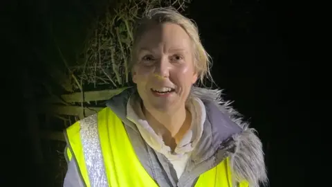 Head and shoulders of Claire Adamson standing in the dark in a high vis vest and grey coat with furry hood. She is smiling and has long blonde hair tied back