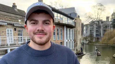Young man with brown hair and beard wearing cap and blue jumper outdoors in front of a building