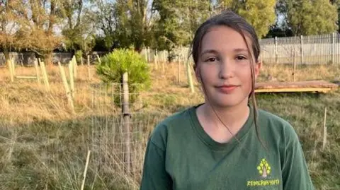 A 12-year-old girl stands in a grassy meadow in front of rows of saplings, with mature trees in the background. She has dark hair, tied back, and wears a green shirt featuring a tree logo and the words "Rewilding Youth".