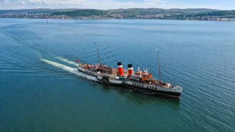 An aerial view of the Waverley as it cruises across a calm sea surface leaving a wake behind it. The boat has a white and black hull and two red, white and black funnels. 
