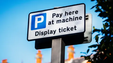 A blue white and black Pay and Display car park sign, set against the backdrop of a blue sky.