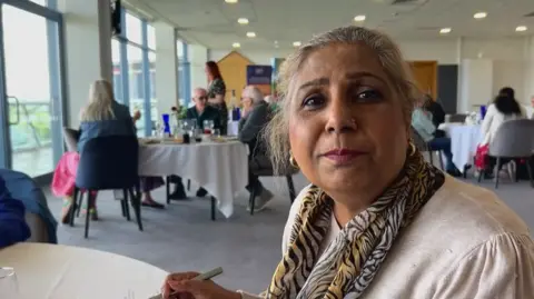 A woman wearing a tiger print scarf and small hoop earrings looks to the camera as she eats a meal