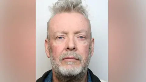 A police mugshot of a man - Sean Morris from Massinger Walk in Swindon - with grey/blue eyes, grey hair, moustache and beard and wearing a black jacket looking directly into the camera. 