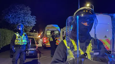 PA Police officers shown on an Ely street holding riot shields and wearing police helmets. Two police vans can be seen in the background. 