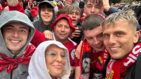 Jack Trotter Mr. Trotter and Friends in the parade. There are eight people in the image. They carry several scarves, hats and Liverpool FC tops. They are smiling and standing for each other.