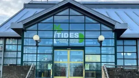 The outside of a leisure centre - the walls are glass and there's a sign saying Tides Leisure and Indoor Tennis Centre in white