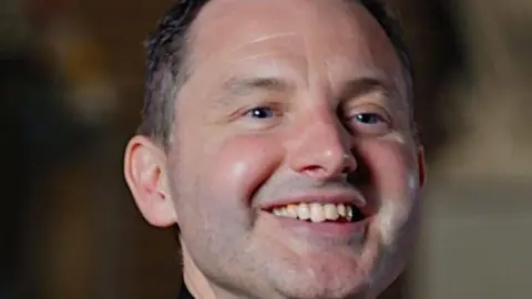 Diocese of Salford Father Frankie Mulgrew is a middle-aged man with short dark hair which is greying at the sides. He is looking off to the left slightly and is smiling broadly. 