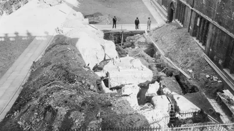 OAHS Oxford Archaeological and Historical Society The remains of Oxford's boundary among rubble and building works in a black and white 19th Century photo. Men in Victorian clothes and hats are visible, one with a cane.