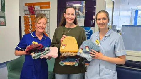 Three women smiling for the camera. Two of the women are wearing hospital nurse uniforms. All three are carrying colourful crocheted beanies.
