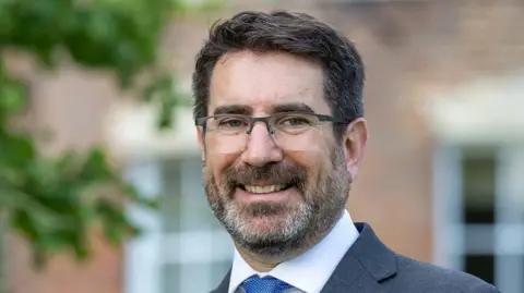 Martyn Poynor David Morton looks ahead smiling in a grey suit with a blue spotty tie and white shirt. He has short black hair and glasses.