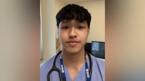 Namling Limbu A young male doctor wearing a purple uniform with a stethoscope around his neck. He is looking into the camera