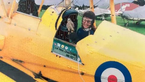 Submitted Tizi Hodson smiles from the pilot's seat of a yellow biplane with an RAF roundel on the side. She is wearing goggles and a pilot's helmet. The plane is parked at an airfield with other planes and a hanger in the background.