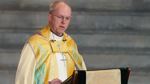 PA Justin Welby wearing glasses, yellow robes and a large silver cross as he sits at a lectern 