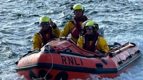 Wells RNLI Three RNLI crew members dressed in yellow waterproof uniforms and helmets. They are on an orange RNLI lifeboat.