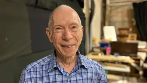 An man with cropped white hair looking towards the camera in a workshop or backstage area. He has a blue chequered shirt on.
