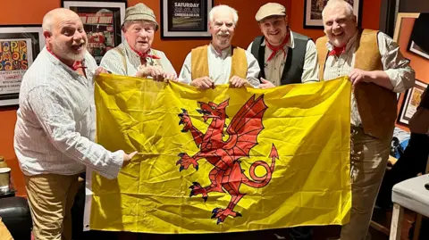 The band standing together behind a Somerset county flag all smiling at the camera.