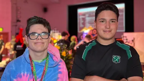 Zach and Ronaldo standing side by side indoors at an event; Zach wears glasses and a colourful hoodie, Ronaldo wears a black sports T‑shirt, with go‑kart displays and stage lighting behind them
