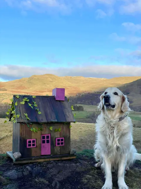 Fiona Gold Golden Labrador dog sitting next to a small wooden dollhouse with hills in the distance