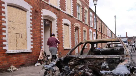 PA Media A burned-out car sits outside a row of boarded-up brick houses as a person walks into a damaged home.