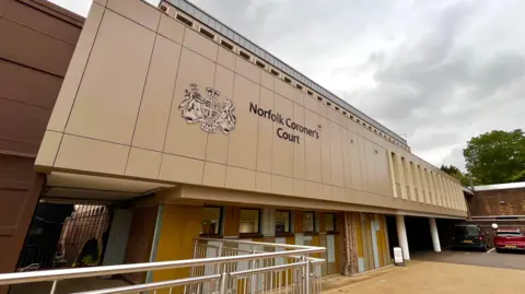 The front of Norfolk Coroner's Court. The building is brown, and has a big logo and title on the front. At the bottom, you can see railings and a walk way. On the right, two cars are parked. 