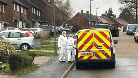 Adrian Harms/BBC Two officers in forensic suits at a van in Hazel Avenue, Guildford. They are both standing near the van, which is red and yellow at the back.