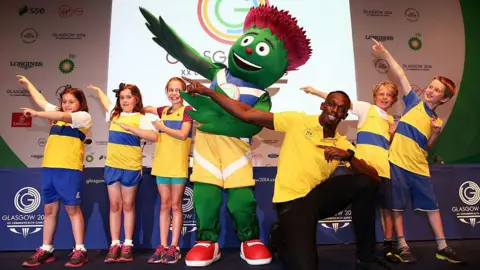 Getty Images Clyde the green and purple animated thistle with his arms outstreched to the left, next to him is sprinter Usain Bolt who is wearing a yellow polo shirt and black trousers. They are surrounded by children and are all doing the same arm gesture- known as Bolt's signature move.
