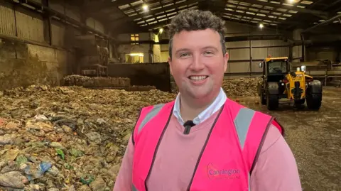 A man smiling in a pink jumper and fluorescent jacket that says 'Cannington Enterprises Ltd' on it. He is standing in a large shed which has what looks like bags of food waste in large piles behind him. There is also a yellow digger behind him.
