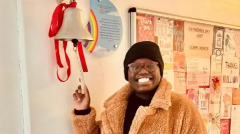 A woman smiling at the camera. She is wearing a black hat and brown fluffy coat. She is ringing the 'remission' bell in the hospital.