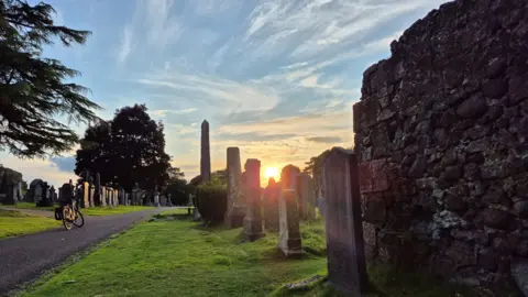 James Kerr A cemetery during golden hour. Several dark-coloured gravestones run up the middle of the image. The sun can be seen going down between the middle two. The sky around it is orange and the rest of the sky is light blue and streaked with white cloud. Two areas of green grass are separated by a grey path running through it, where a bike is parked.
