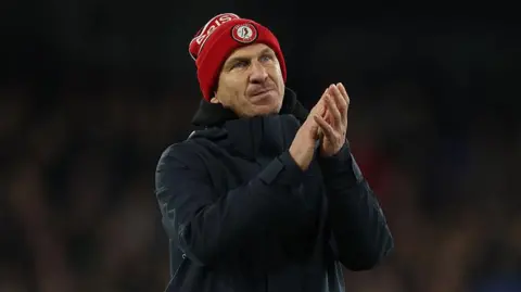 Gerhard Struber wearing a Bristol City bobble hat and coat and clapping his hands while walking on the pitch at the end of the match with Ipswich