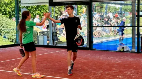 De La Mare family Two people are on a red clay padel court, giving each other a high-five. One person is wearing a green shirt and black shorts, while the other is dressed in black sportswear. Both are holding padel rackets. In the background, there is an audience watching through the glass walls of the court, with some greenery and another player visible outside.