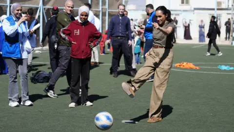 BBC Food La duquesa de Sussex patea una pelota mientras las niñas miran desde el interior de una cancha de césped artificial.
