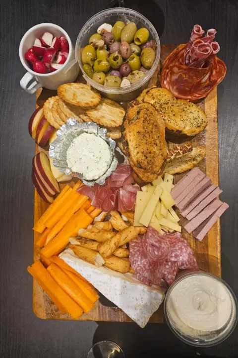 Jon Rowland-Beer A charcuterie board full of salami, olives, cheese swirls, hummus dip, slices of different cheeses and carrots. The photo has been taken from above to showcase all of the food on display.