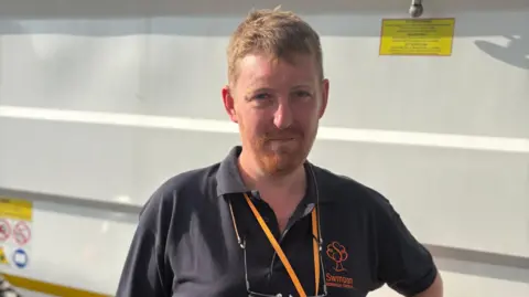 A ginger-haired man standing in front of a white recycling truck wearing an orange lanyard and a dark polo shirt with a Swindon Borough Council logo on it.