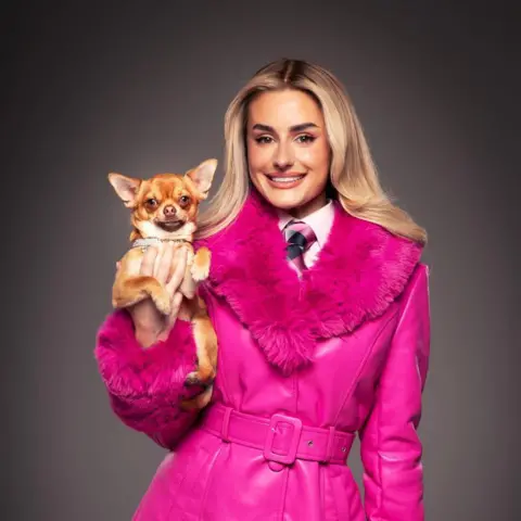 Matt Crockett Amber Davies dressed as the character Elle Woods. She has blonde hair, wears a bright pink coat with a furry trim, and holds a chihuahua.