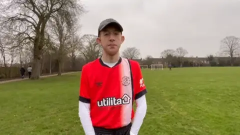 Family photo Aidan wearing a red and white Luton Town football shirt looking at the camera. He is standing on a football pitch.