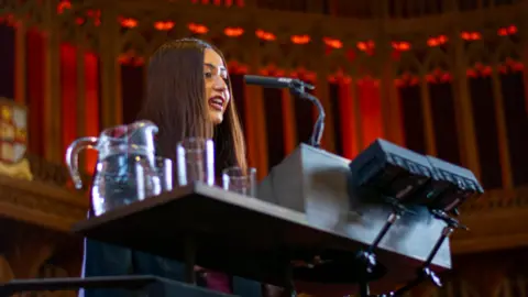 University of Bristol Bristol University masters student Gurvin Chopra is standing behind a lectern speaking into a microphone. She has long brown hair.