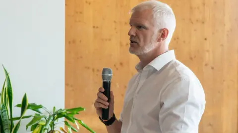 A side profile of a man with cropped white hair and close-cut beard addressing a room, holding a microphone. The man is wearing a white shirt and standing by a wooden panel background.