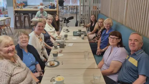 Stella Cleary A group of eleven people sitting around a row of tables in a cafe looking at the camera and smiling. There is a variety of food and drink on the table. They are sitting on a blue leather booth bench with light wooden panelling behind.