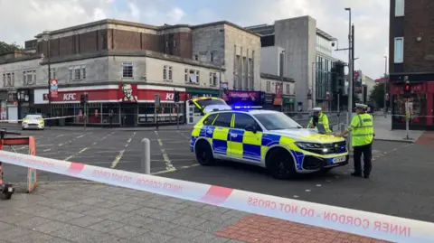 Police cordon with two police cars inside the cordon at a large city centre junction.  Two police officers stand at police car seen to the right.