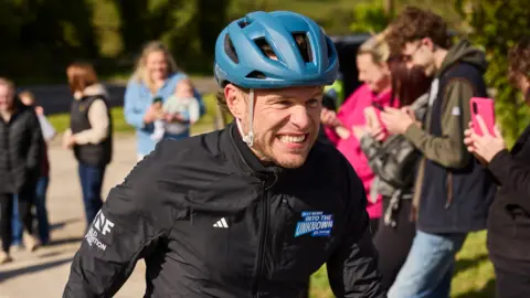 Olly Murs in a blue cycle helmet and black jacket with a grimace on his face as he takes part in the challenge. A crowd of supporters are behind him.