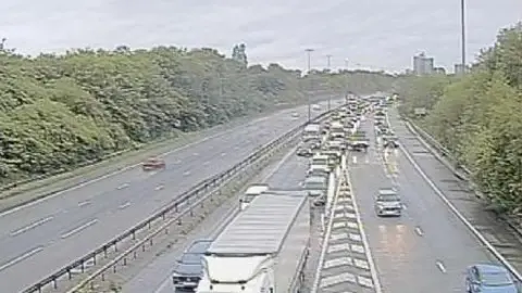 A CCTV image showing a line of traffic on a motorway, surrounded by trees. 